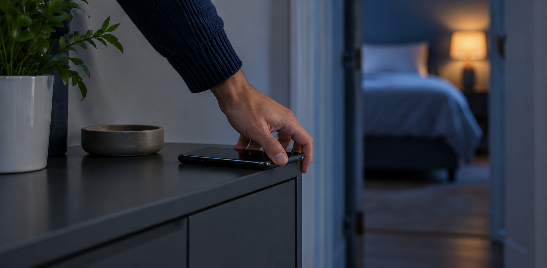 A hand leaving a phone on a dresser outside a softly lit bedroom.